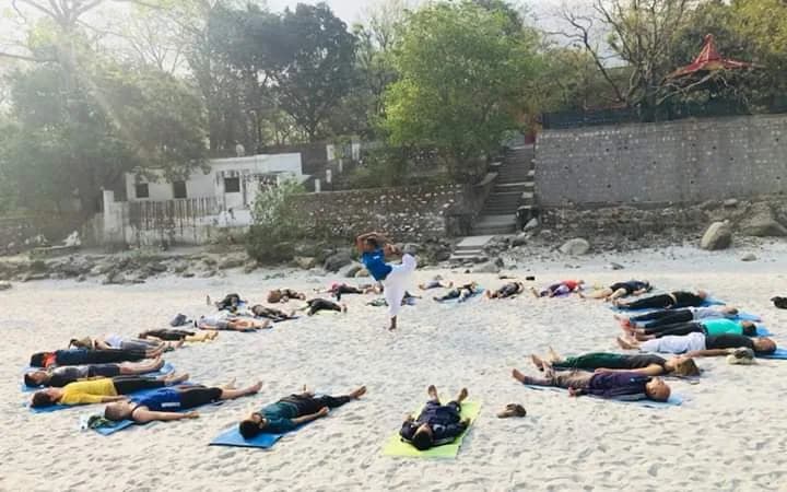 Deep Yoga cohort in savasana on the Ganges beach at Tapovan, Rishikesh, with the lead teacher demonstrating a posture